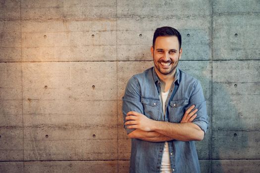 Young successful smiling entrepreneur standing in front of a concrete wall with crossed hands and looking at camera.