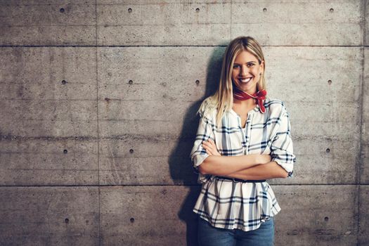 Young beautiful successful smiling female entrepreneur standing in front of a concrete wall with crossed hands and looking at camera.