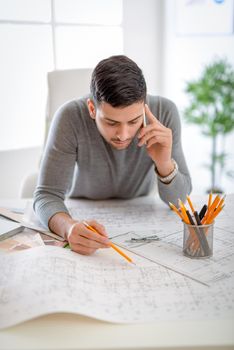 Young arabian male constructor analyzing blueprint at desk in office and using phone.