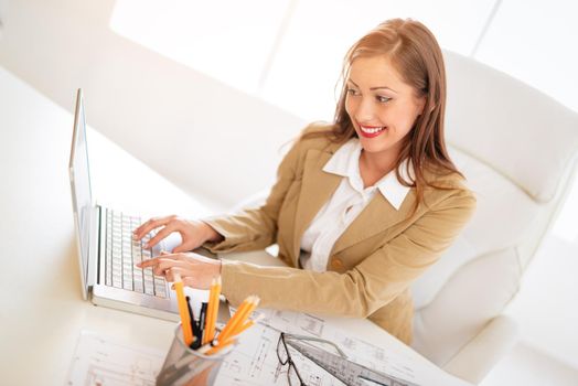 Smiling young woman architect working on laptop at desk in office.