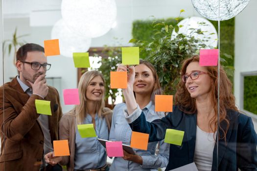 Creative professionals standing and discussing at the office behind glass wall with sticky notes and looking a post it note wall.
