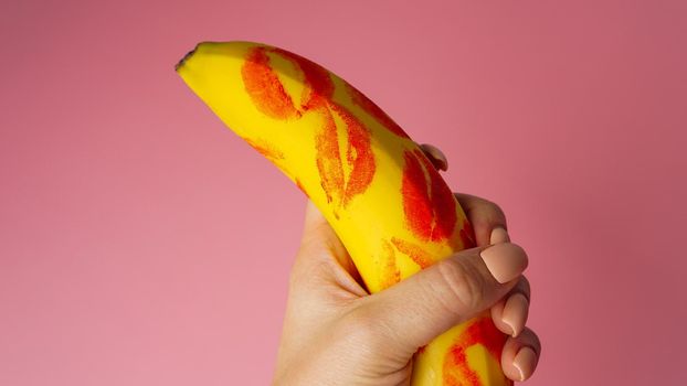 Woman hand holding banana with red lipstick markings on a pink background