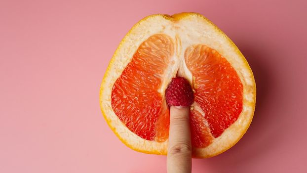 Vagina symbol. Finger with raspberry on grapefruit on pink background. Sex concept.