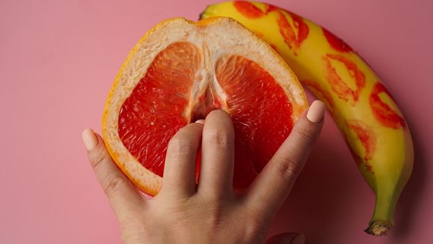 Fingers in grapefruit on pink background. Composition with fresh banana with traces of red lipstick and grapefruit on pink background.