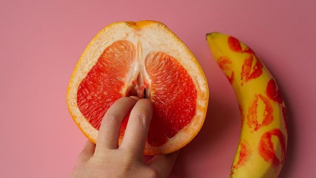 Fingers in grapefruit on pink background. Composition with fresh banana with traces of red lipstick and grapefruit on pink background.