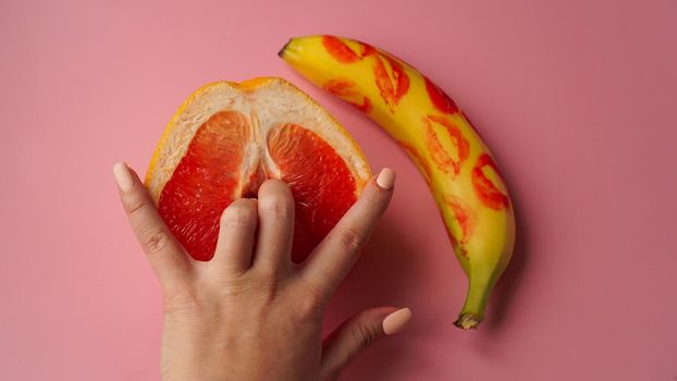 Fingers in grapefruit on pink background. Composition with fresh banana with traces of red lipstick and grapefruit on pink background.