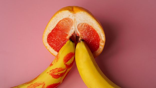 Two bananas and grapefruit on a pink background. There are traces of red lipstick on the banana