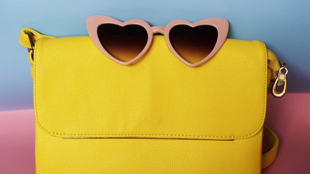 Yellow bag and heart-shaped sunglasses on pink and blue background - summer picture