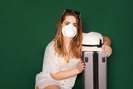 Beautiful girl in a white summer dress with a suitcase. Medical mask on the face. Closed borders during coronavirus. Tourist on a green background