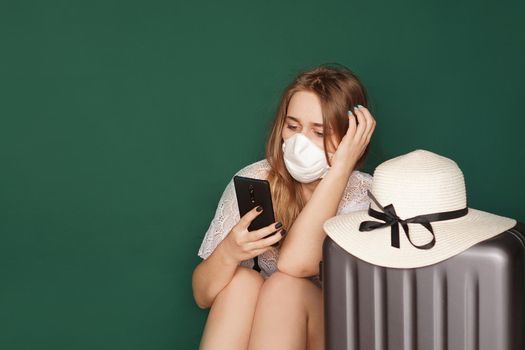 Girl tourist in a medical mask sits with luggage, phone in hands, on a green background. COVID-19 outbreak of coronavirus. Travel concept