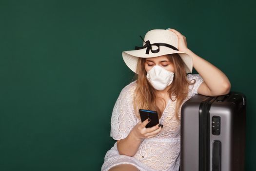 Girl tourist in a medical mask sits with luggage, phone in hands, on a green background. COVID-19 outbreak of coronavirus. Travel concept