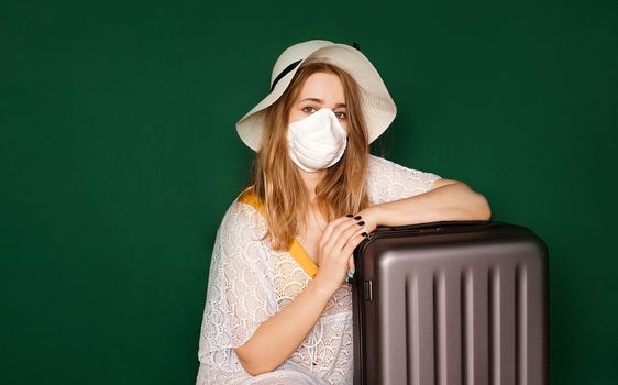 Beautiful girl in a white summer dress with a suitcase. Medical mask on the face. Closed borders during coronavirus. Tourist on a green background