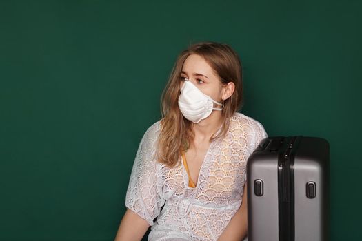 Beautiful girl in a white summer dress with a suitcase. Medical mask on the face. Closed borders during coronavirus. Tourist on a green background