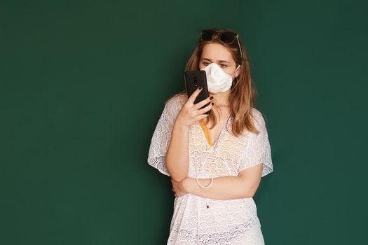 Girl tourist in a medical mask, phone in hands, on a green background. COVID-19 outbreak of coronavirus. Travel concept