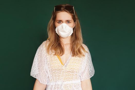 Beautiful girl in a white summer dress. Medical mask on the face. Closed borders during coronavirus. Tourist on a green background