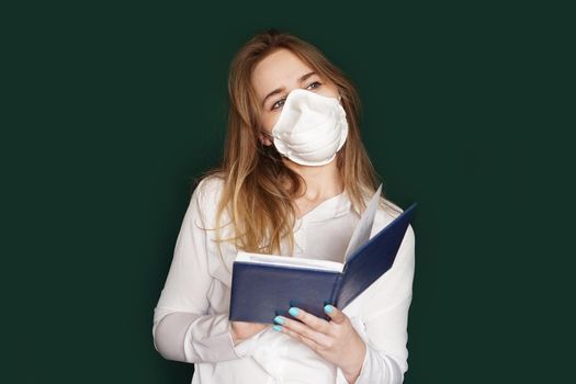 Young girl in a medical mask in a white office blouse with a notebook in hand on a green background