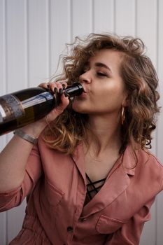 A young woman with curly hair drinks from a bottle of champagne. She is indoors on a white background