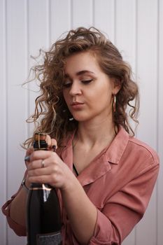A young woman with curly hair opens a bottle of champagne. She is indoors on a white background