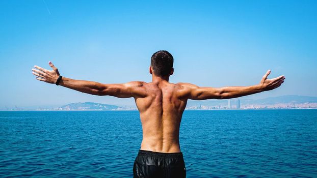 Man Raising His Hands or Open arms standing back looking to sea blue sky horizon. Strong muscular Men, perfect body, arms, back. Freedom Concept