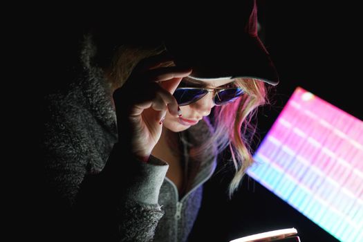 Hacker girl in a cap and sunglasses in front of a computer in neon light holds smartphone in hand