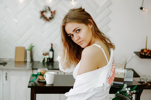 A girl in a long white shirt stands in the kitchen. The white kitchen is decorated with Christmas decorations and garlands