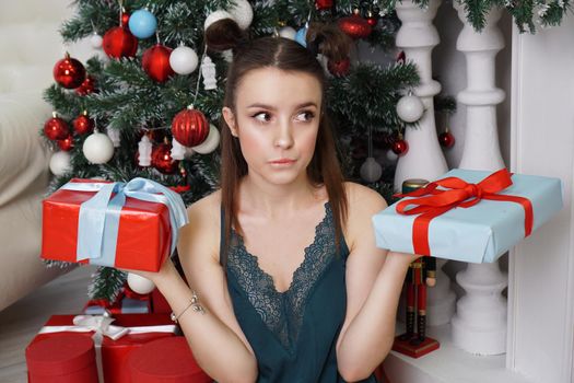 Happy girl in a green peignoir and funny hairstyle holding two gift boxes, not knowing which gift to choose to give a Christmas tree, decorated room
