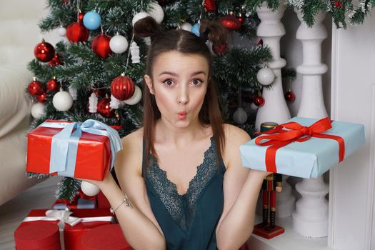 Happy girl in a green peignoir and funny hairstyle holding two gift boxes, not knowing which gift to choose to give a Christmas tree, decorated room