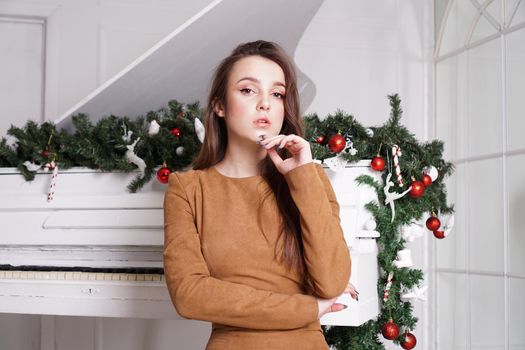 Beautiful sensual brunette girl with long straight hair near a white piano with Christmas decoration