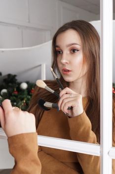 Makeup artist with long dark hair holding a powder brush, haughty expression, reflection in the mirror, christmas decor behind