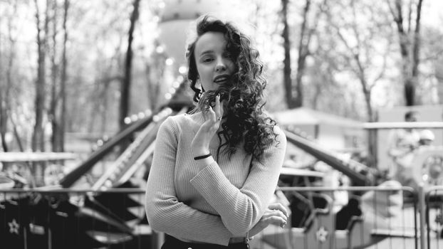 Girl chilling in amusement park in weekend morning. Laughing good-humoured female model with curly hair standing near carousel. Black and white photo