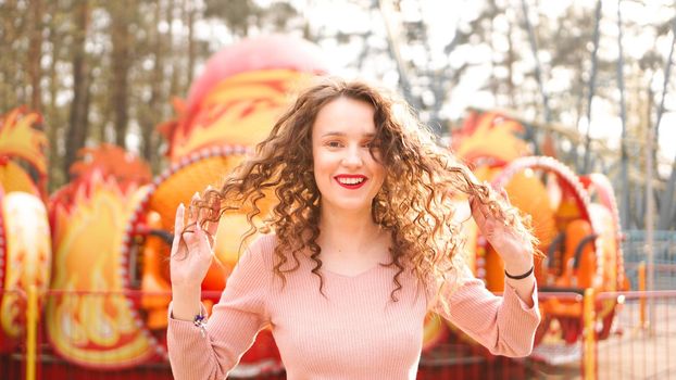 Girl chilling in amusement park in weekend morning. Laughing good-humoured female model with curly hair standing near carousel.