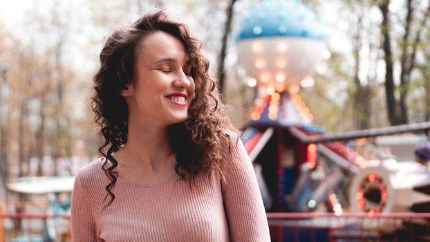 Girl chilling in amusement park in weekend morning. Laughing good-humoured female model with curly hair standing near carousel.