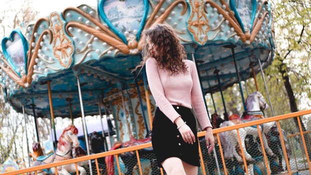 Girl chilling in amusement park in weekend morning. Laughing good-humoured female model with curly hair standing near carousel.