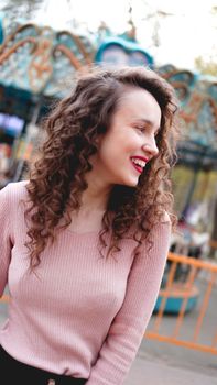 Girl chilling in amusement park in weekend morning. Laughing good-humoured female model with curly hair standing near carousel.