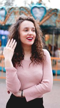 Girl chilling in amusement park in weekend morning. Laughing good-humoured female model with curly hair standing near carousel.