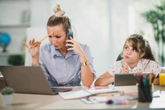 Busy single mother using laptop and talking on her smartphone while her cute daughter learning at home.