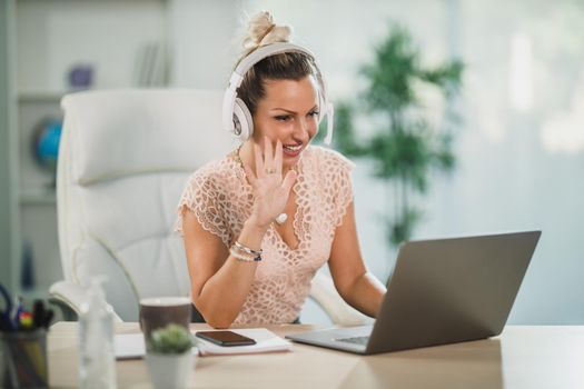 An attractive business woman working in a call center and waving to client over a video call at her laptop.