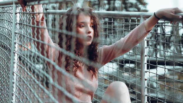 Fashion portrait of trendy young woman sitting near the netting rabitz in the city