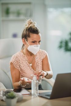 An attractive young businesswoman with N95 protective mask using antiseptic gel to disinfect her hands at her home office during corona virus pandemic.