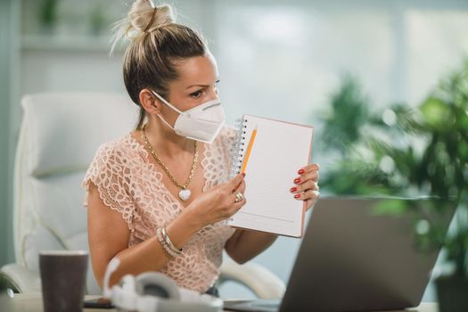An attractive business woman with N95 protective mask working in a call center and talking with client online at her laptop.