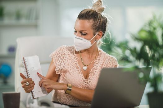 An attractive business woman with N95 protective mask sitting alone in her home office and working online at her laptop.