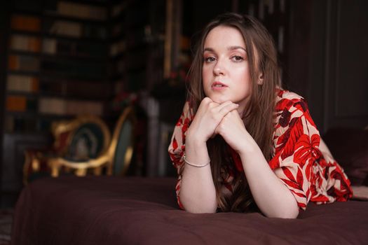 Relaxed woman lying on a brown bed thinking deeply. Woman in retro style room in red dress