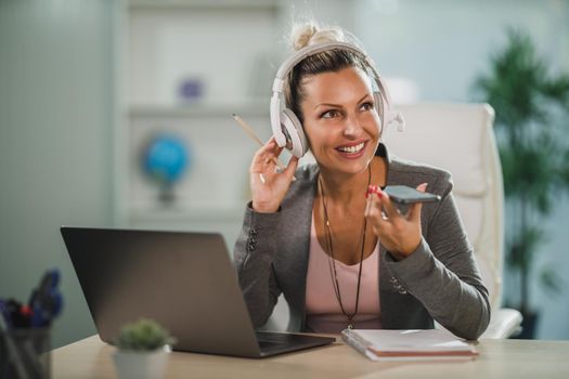 An attractive business woman sending voice message on cellphone while working online at laptop in her home office.