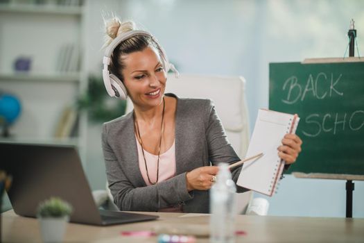 A smiling young woman making video call at her laptop and teaching students online in her home office.