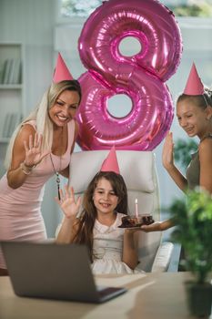 Beautiful young mom with her cute daughters have video call with friends while birthday celebration at home during pandemic isolation.