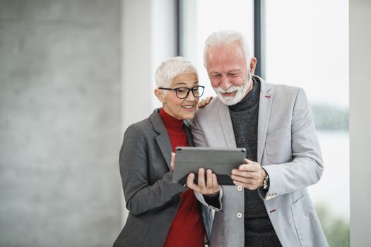 A successful senior businessman and his smiling female colleague making video call on a digital tablet in a modern office.