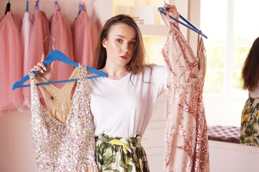 Young woman standing near wardrobe, holding dress on hangers, trying to decide what to wear. Pretty female choosing clothes in dressing room.