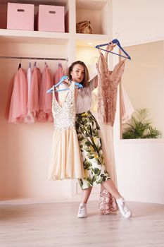 Young woman standing near wardrobe, holding dress on hangers, trying to decide what to wear. Pretty female choosing clothes in dressing room.