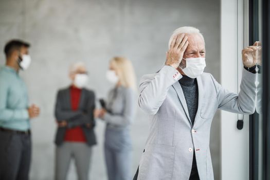A tired senior businessman with protective mask standing in a modern office and looking out the window during corona virus or COVID-19 pandemic.