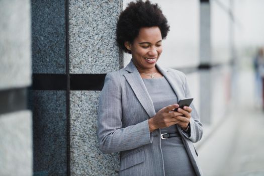 Portrait of a smiling black business woman typing textual message on a smart phone during quick break in front a corporate building.